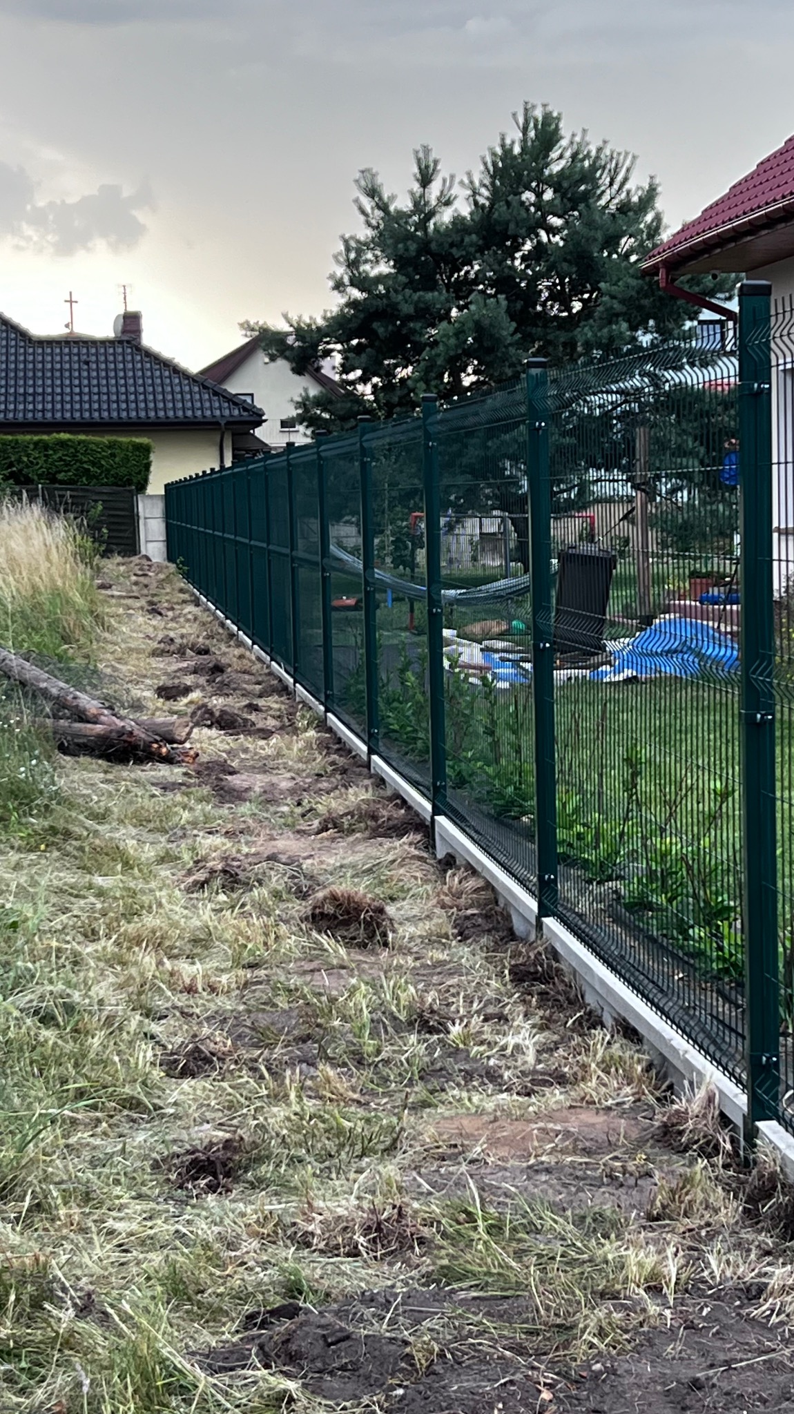 Długi, ciemnozielony płot z paneli z betonową podmurówką, widziany z boku, z widocznymi śladami po wykopach w trawie. W tle domy i drzewa pod pochmurnym niebem.
