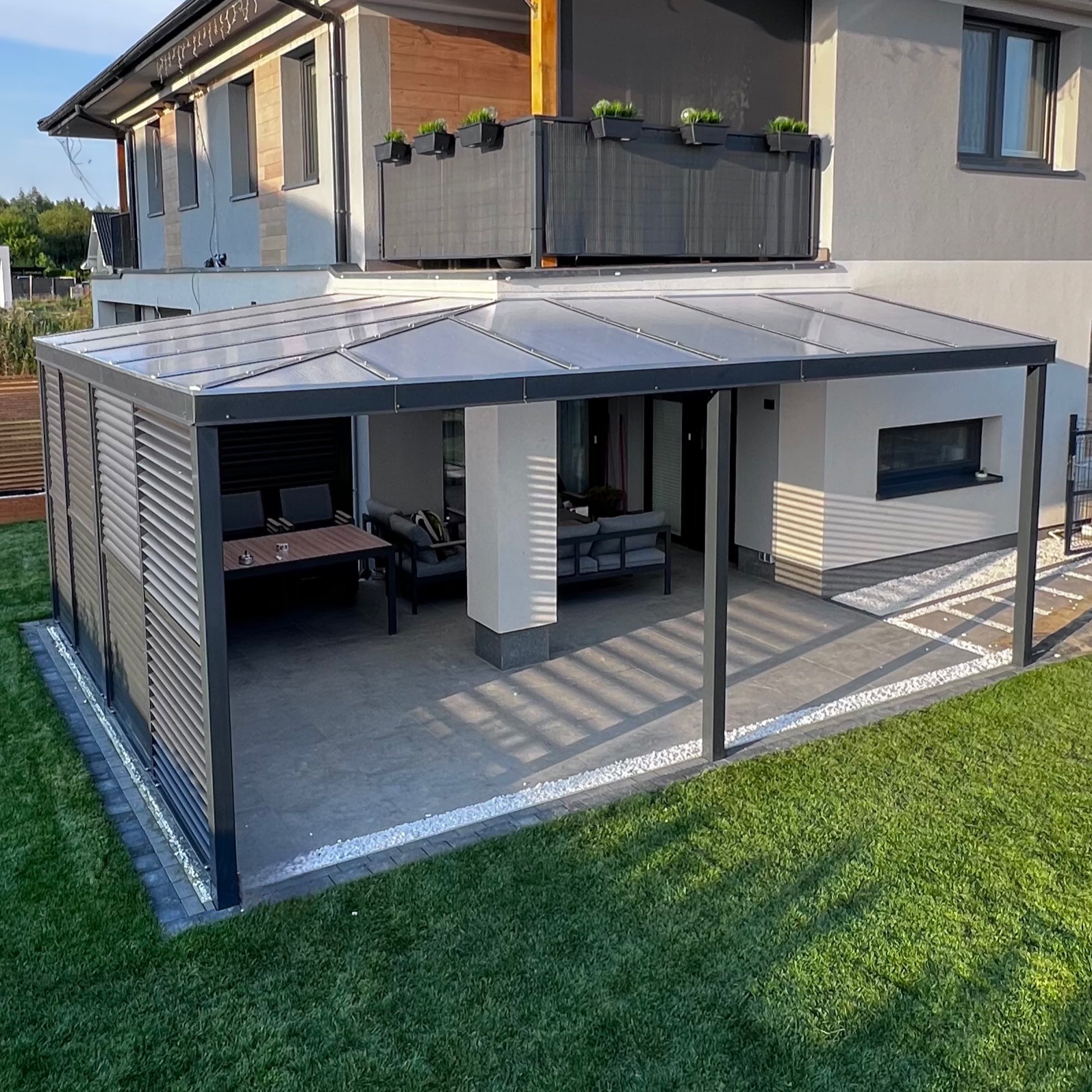 Nowoczesna pergola z aluminiową konstrukcją i przezroczystym zadaszeniem, przylegająca do domu, z meblami ogrodowymi wewnątrz i dekoracyjnym obramowaniem z białych kamieni.