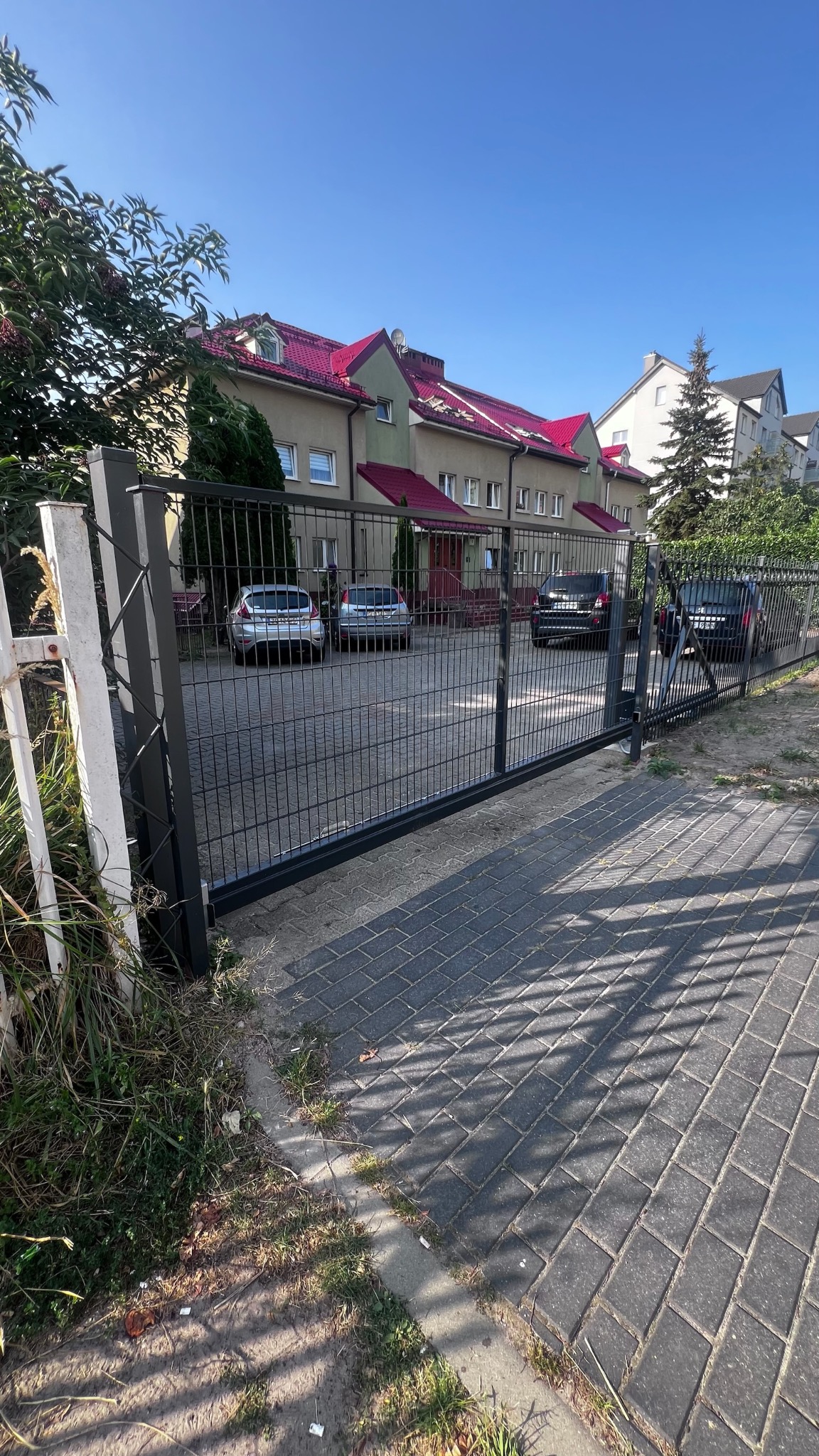 Czarna brama wjazdowa panelowa z widokiem na parking przed budynkiem z czerwonym dachem w słoneczny dzień.