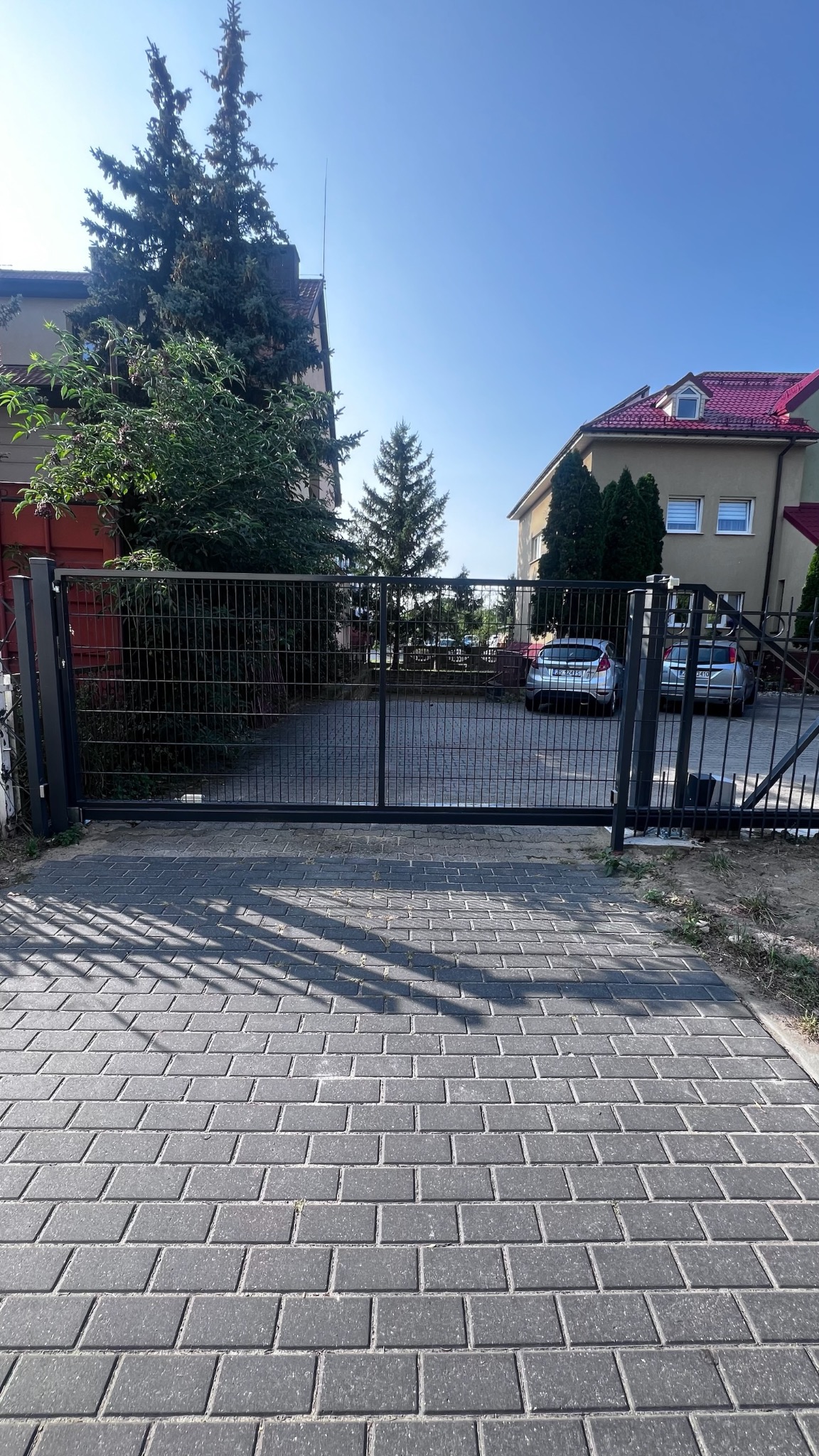 Zewnętrzna brama przesuwna z panelowym wypełnieniem, widoczna z zewnątrz posesji, z szarej kostki brukowej, w tle dwa zaparkowane samochody i budynki mieszkalne w słoneczny dzień.
