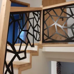 Jasne drewniane schody z nowoczesną, geometryczną, czarną balustradą, widok z dołu na górę, w tle widoczne drzwi i fragment ceglanej ściany.