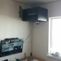 Monitor z podglądem z kamer, szafa rack z otwartymi drzwiczkami na ścianie, kamera kopułkowa zamontowana pod szafą, widoczne narzędzia na biurku.