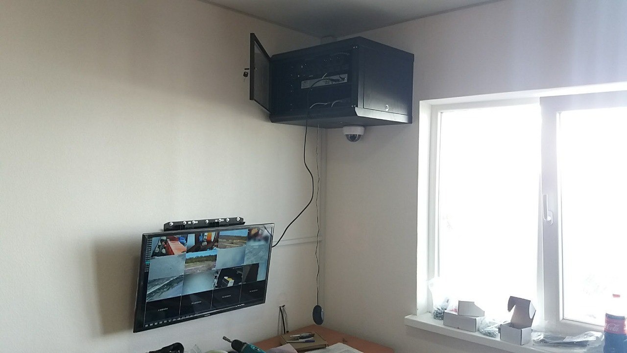 Monitor z podglądem z kamer, szafa rack z otwartymi drzwiczkami na ścianie, kamera kopułkowa zamontowana pod szafą, widoczne narzędzia na biurku.