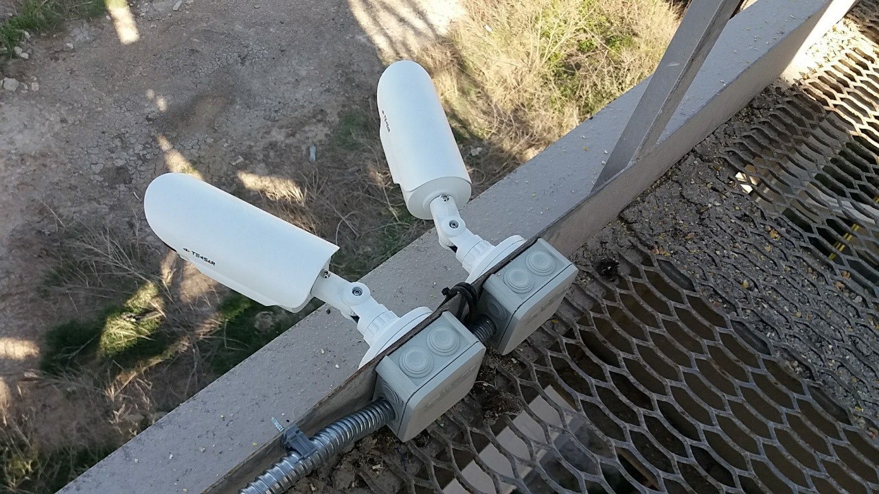 Dwie białe kamery monitoringu zamontowane na metalowej konstrukcji z kratą, z widocznymi puszkami instalacyjnymi i peszlem.