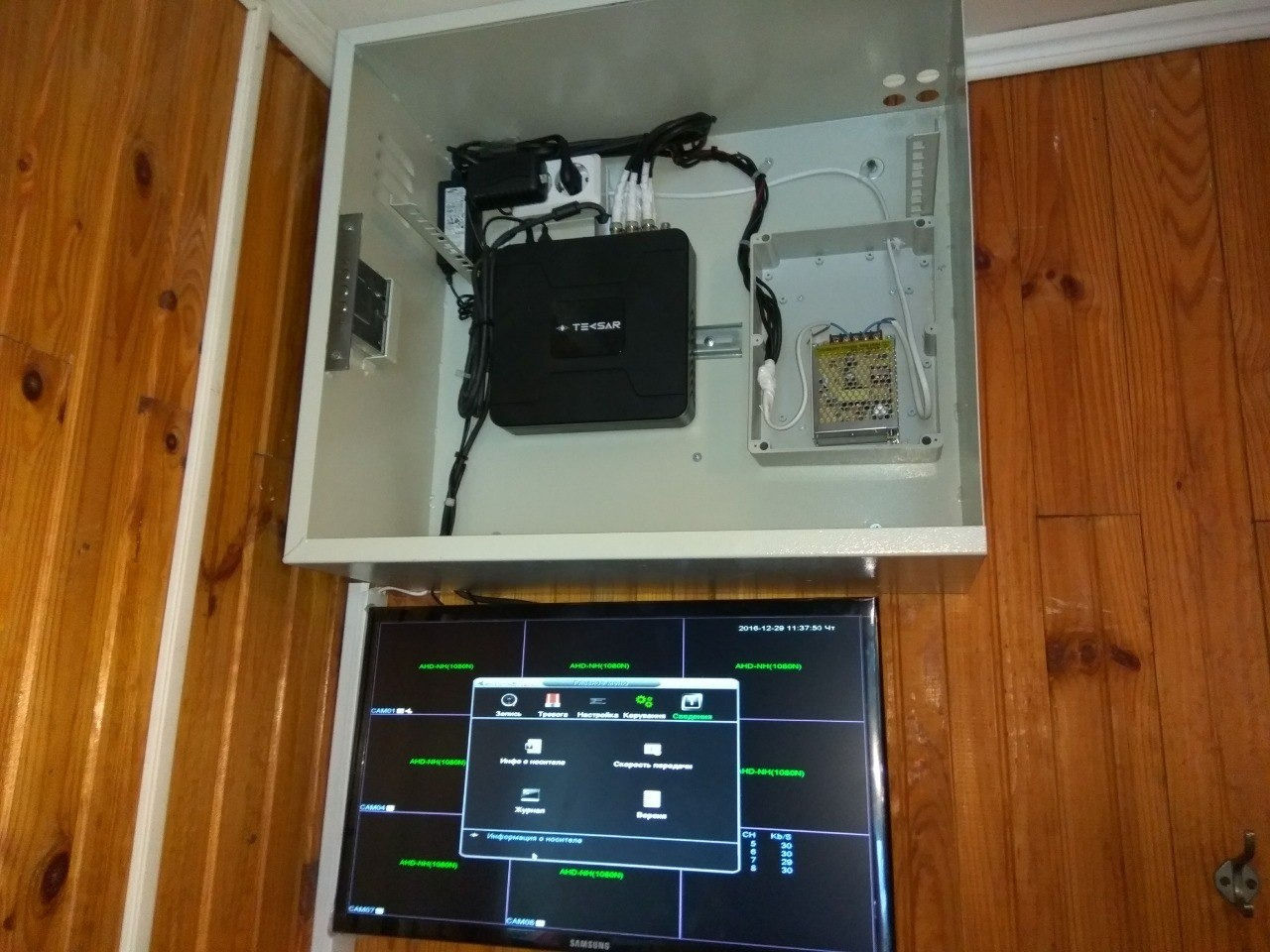 Zamknięta szafka z urządzeniami elektronicznymi, w tym czarna skrzynka z napisem TE SAR, oraz monitor Samsung wyświetlający obraz z systemu kamer, zamontowane na drewnianej ścianie.