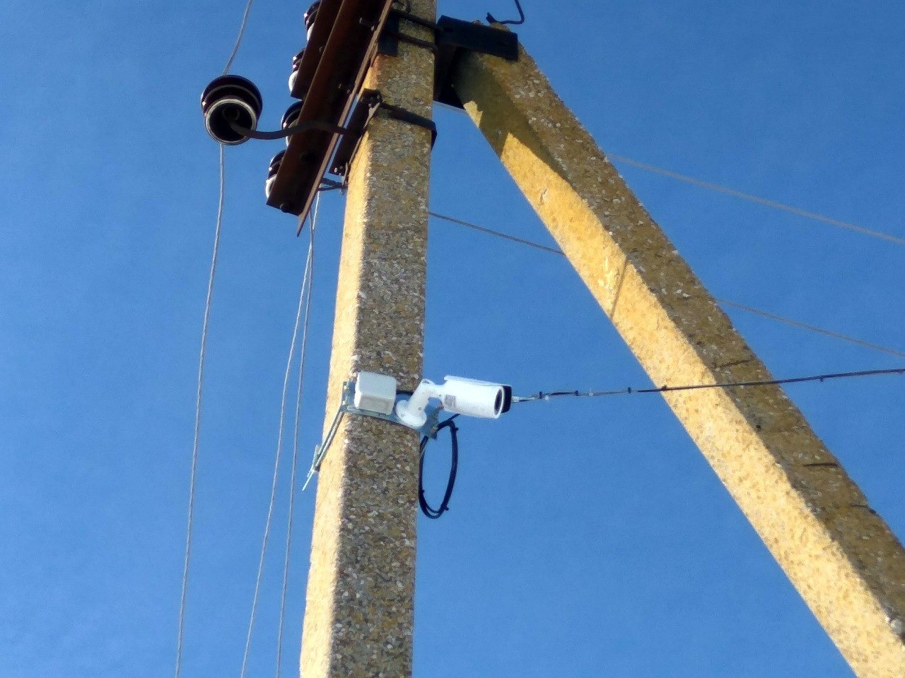 Biała kamera monitoringu zamontowana na betonowym słupie energetycznym z widocznymi przewodami na tle błękitnego nieba.