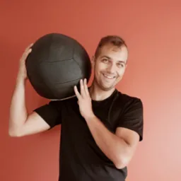 Uśmiechnięty mężczyzna w czarnej koszulce trzyma dużą czarną piłkę lekarską na ramieniu na tle jednolitej, koralowej ściany.