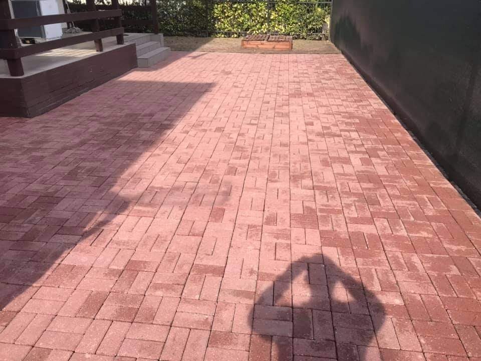 Czerwona kostka brukowa ułożona w jodełkę, z widocznym cieniem osoby fotografującej, przylegająca do zielonego żywopłotu i drewnianego podestu.