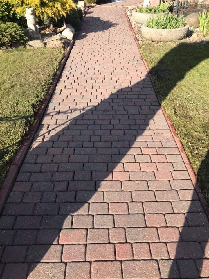 Czerwono-brązowa kostka brukowa na ścieżce w Kołobrzegu, z widocznym cieniem drzewa i zadbanym trawnikiem po bokach.