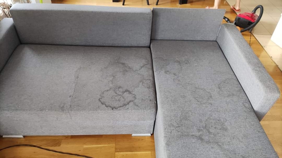 Szara, narożna sofa z widocznymi plamami przed czyszczeniem, obok odkurzacz piorący i fragment stopy w klapku.