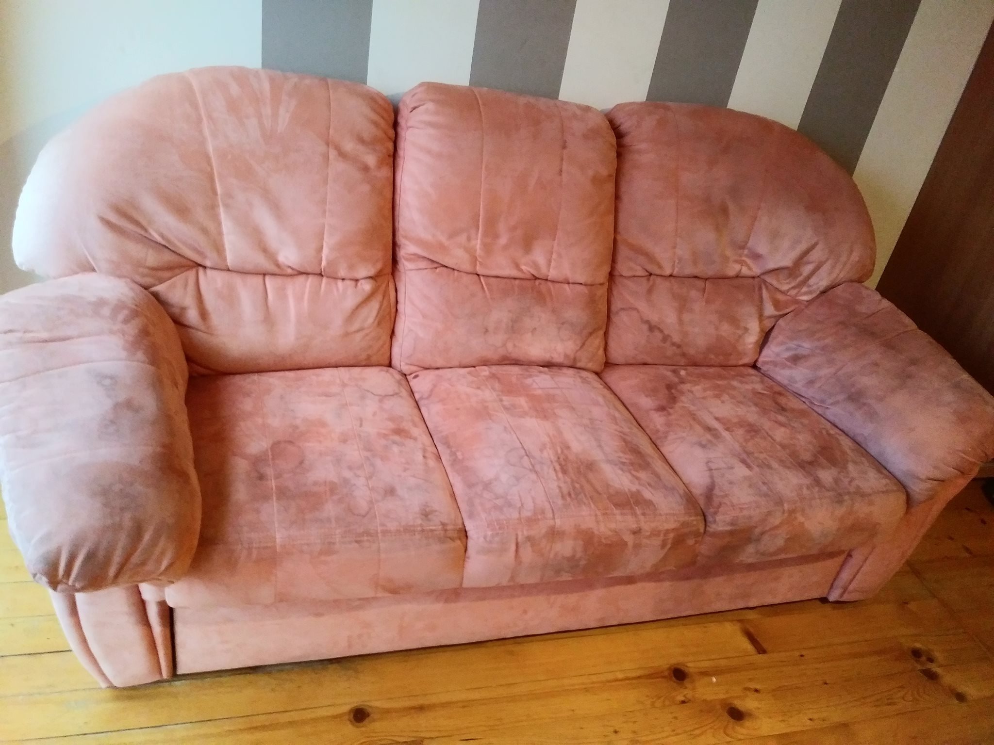 Trzyosobowa, pluszowa kanapa w odcieniach brudnego różu, z widocznymi plamami na siedzisku, ustawiona na drewnianej podłodze przy ścianie w szaro-białe pasy.