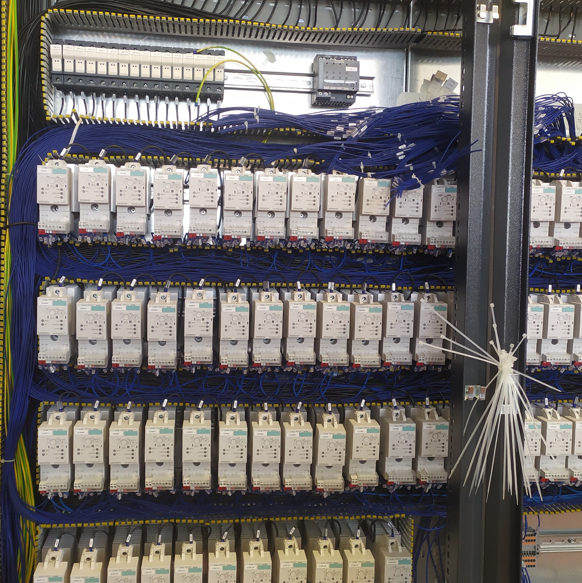 Rozdzielnica elektryczna z licznymi przekaźnikami Siemens, okablowaniem w kolorze niebieskim i żółto-zielonym, oraz wiązką białych opasek zaciskowych przy metalowej konstrukcji.
