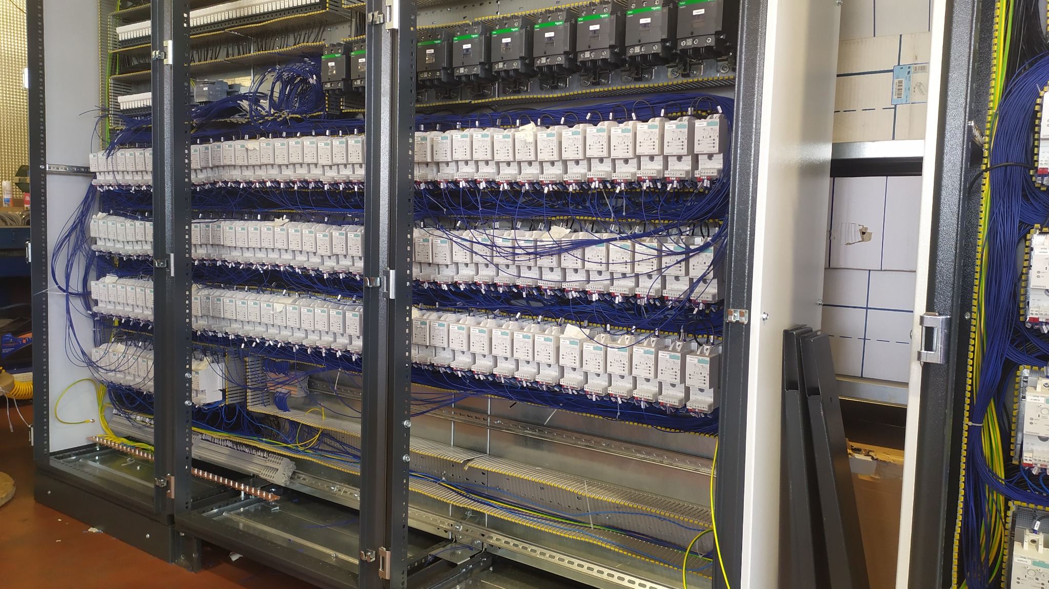 Rozbudowana szafa sterownicza z licznymi modułami, gęsto okablowana niebieskimi przewodami, widoczne szyny montażowe i elementy mocujące, częściowo otwarte drzwi szafy odsłaniające puste kartony.
