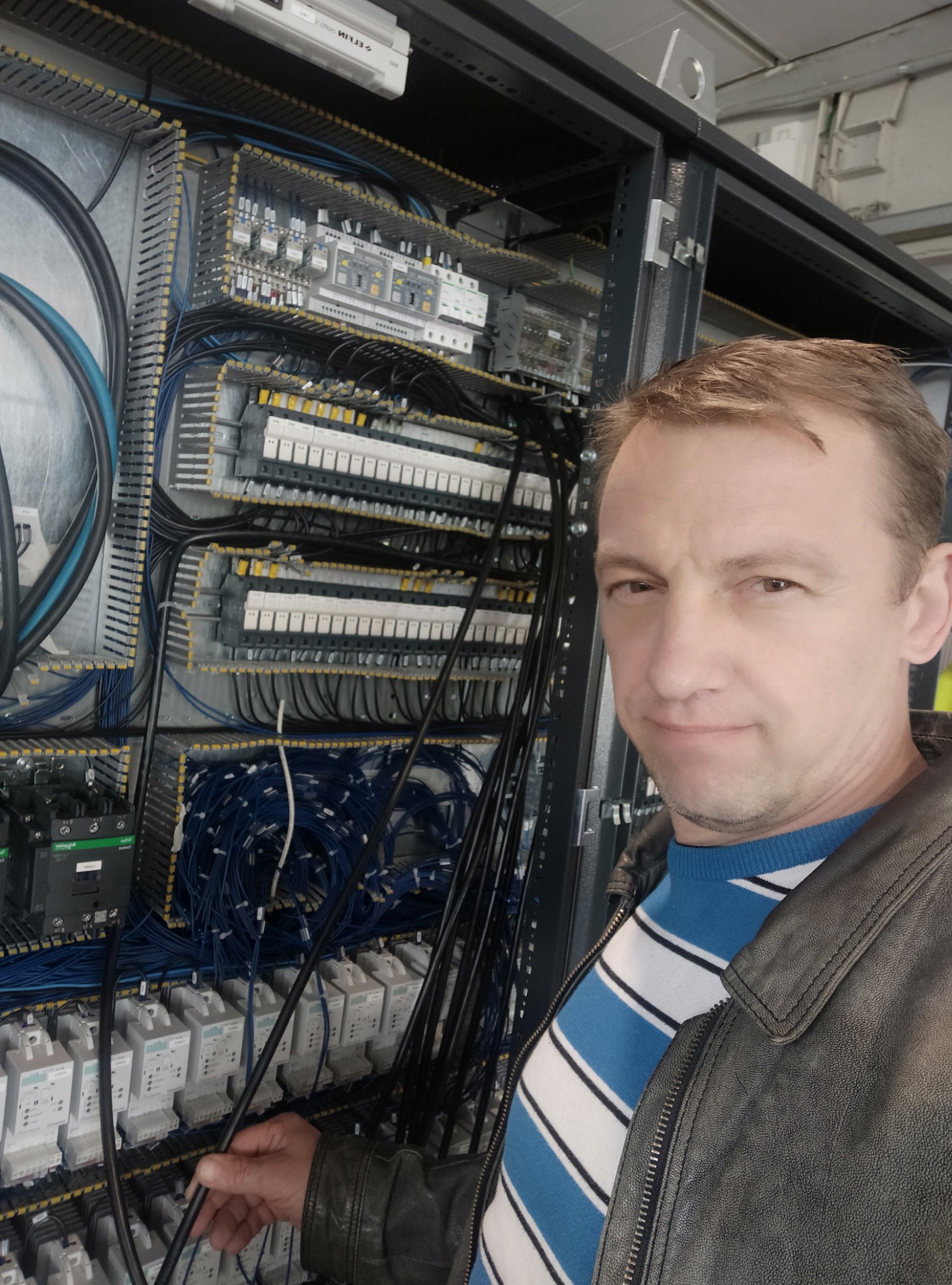 Elektryk w skórzanej kurtce i swetrze w paski, pozujący na tle rozbudowanej szafy sterowniczej z licznymi przewodami, przekaźnikami i listwami zaciskowymi.