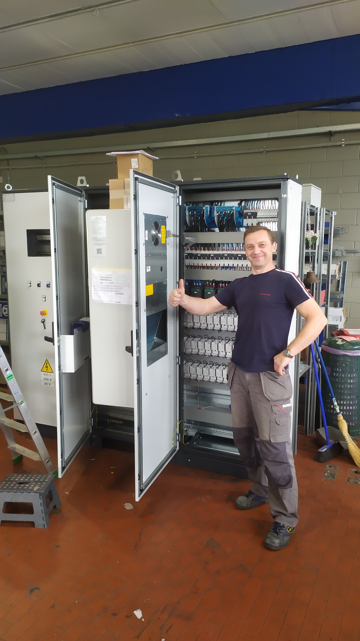 Uśmiechnięty elektryk z podniesionym kciukiem prezentuje rozbudowaną szafę sterowniczą z widocznymi elementami instalacji elektrycznej, w tle drabina i narzędzia na podłodze z terakoty.