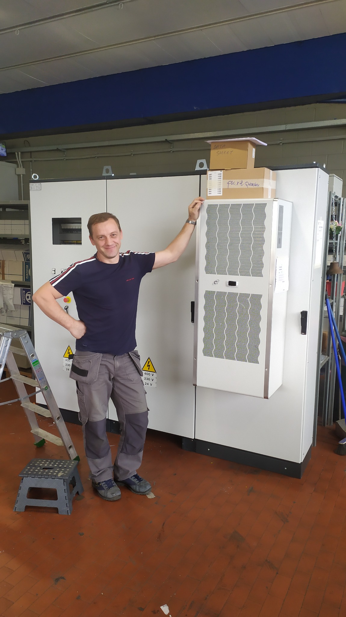 Elektryk w szarych spodniach i granatowej koszulce opiera się o dużą, białą szafę sterowniczą z elementem chłodzącym, obok drabina i stołek, na szafie kartony z dokumentacją.
