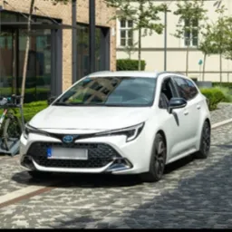 Biała Toyota Corolla kombi zaparkowana na brukowanej ulicy w słoneczny dzień, rower w tle. Nowoczesna architektura i zieleń w otoczeniu.