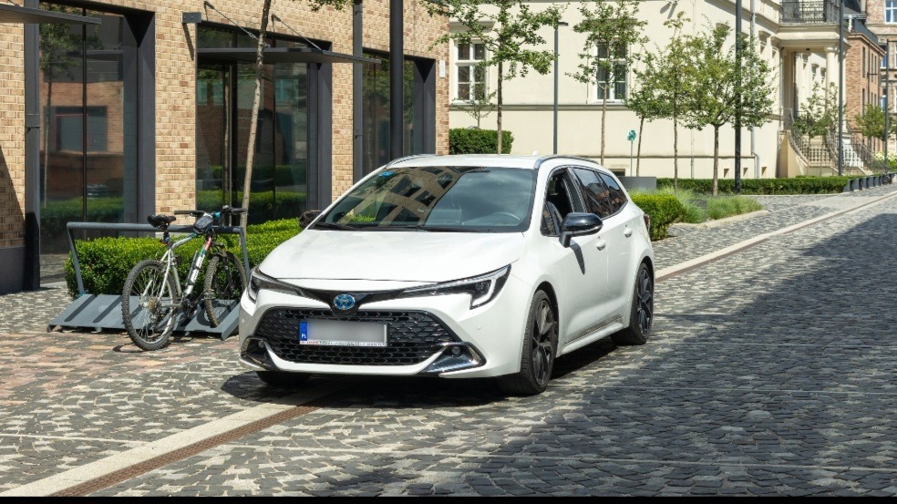 Biała Toyota Corolla kombi zaparkowana na brukowanej ulicy w słoneczny dzień, rower w tle. Nowoczesna architektura i zieleń w otoczeniu.