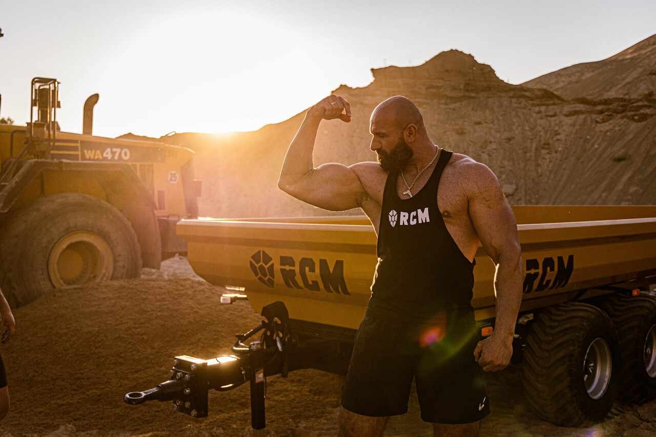 Mężczyzna prezentuje muskulaturę na tle żółtej przyczepy budowlanej z logo RCM i ładowarki WA470 w kamieniołomie o zachodzie słońca.