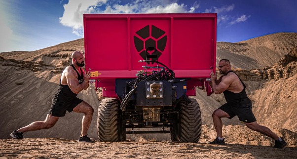 Dwóch barczystych mężczyzn w sportowych strojach napiera na czerwoną przyczepę rolniczą z czarnym logo na tle piaszczystego krajobrazu.