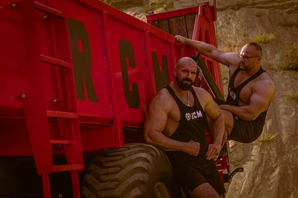 Dwóch muskularnych mężczyzn w czarnych podkoszulkach pozuje przy czerwonej, dużej przyczepie rolniczej z napisem 'RCM'.