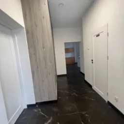 Nowoczesny korytarz z czarną marmurową podłogą i zabudową meblową w odcieniach szarości. Widoczne drzwi i fragment kuchni w tle.