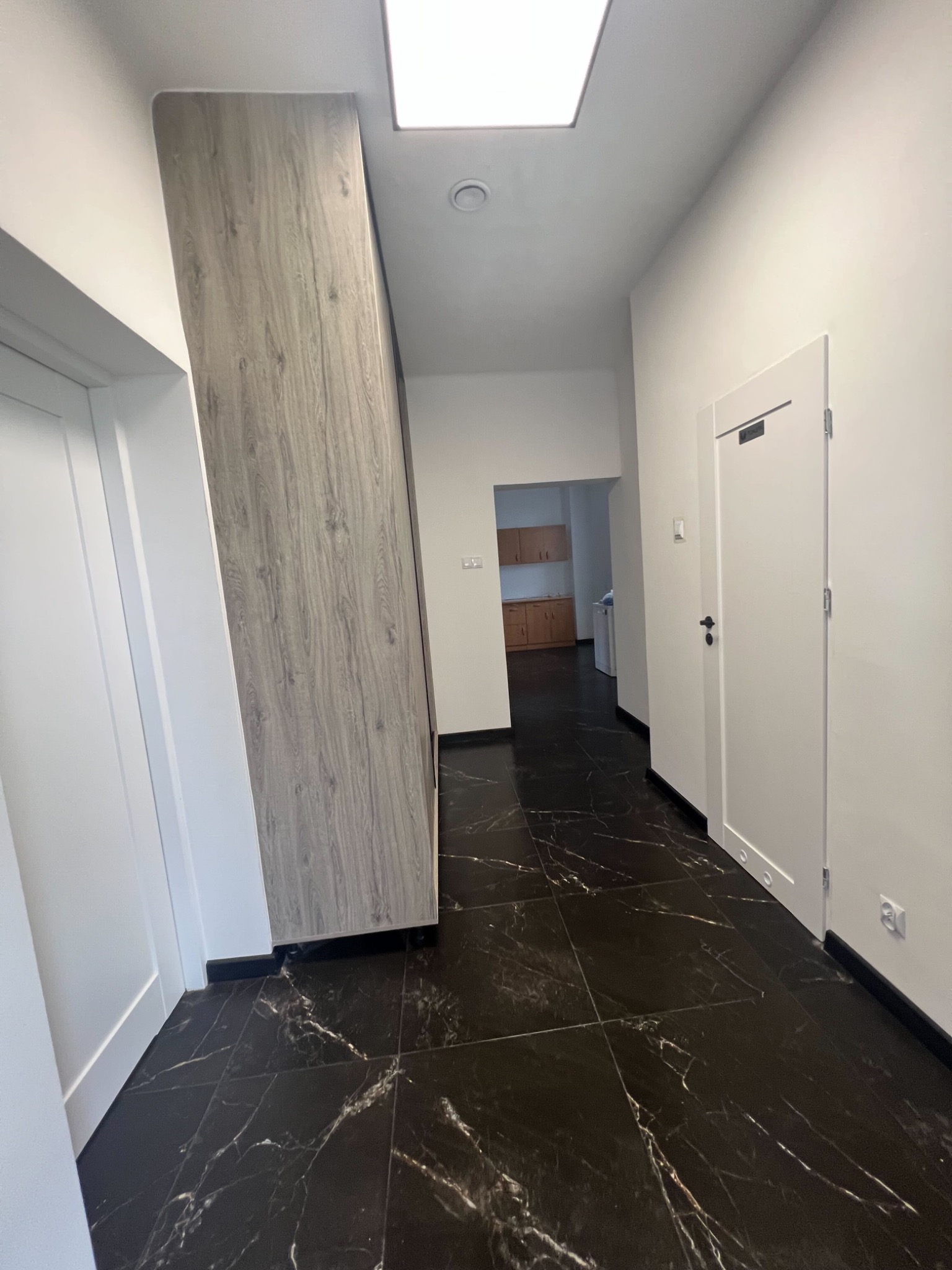 Nowoczesny korytarz z czarną marmurową podłogą i zabudową meblową w odcieniach szarości. Widoczne drzwi i fragment kuchni w tle.