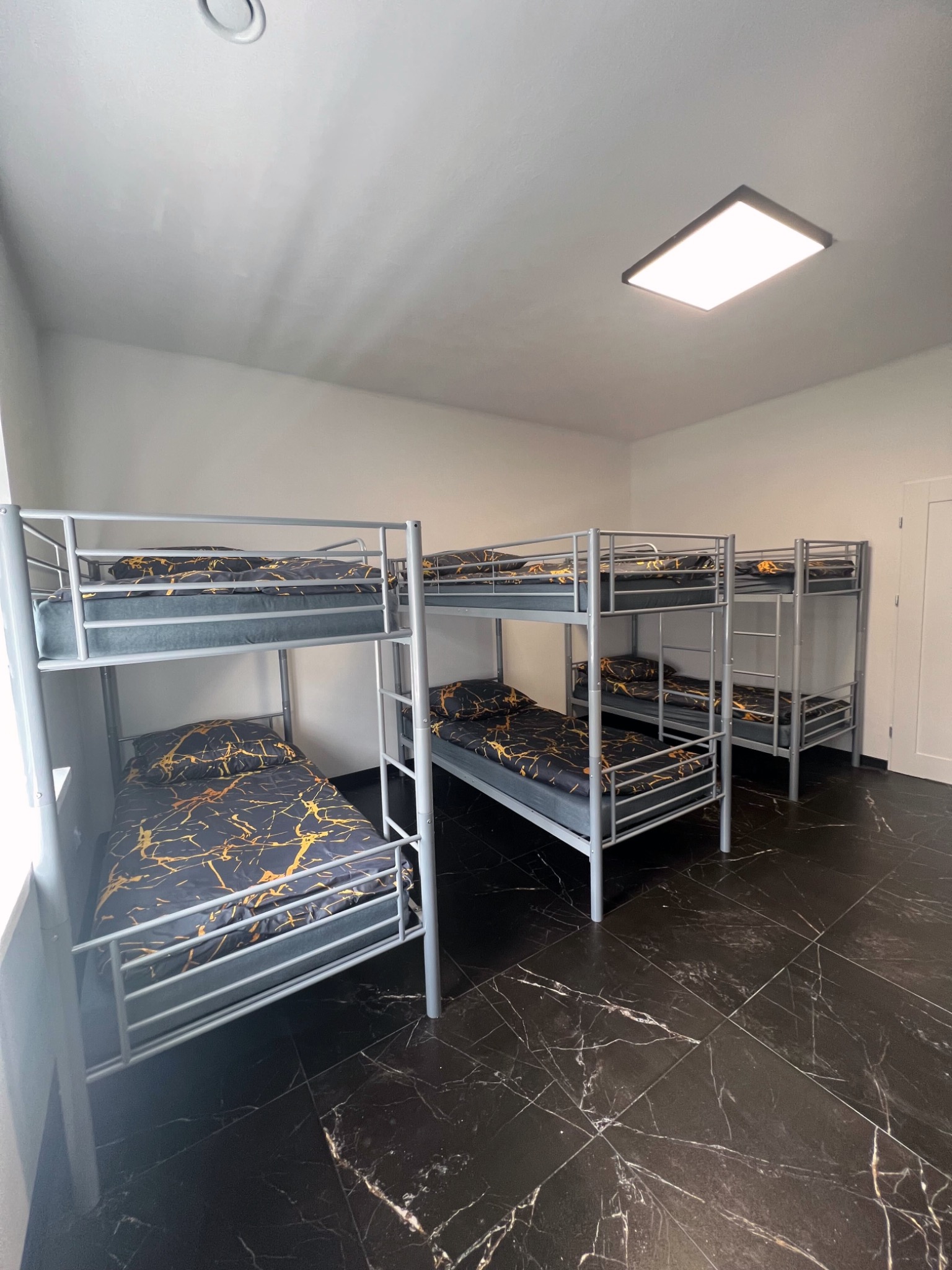 Pokój z trzema łóżkami piętrowymi na ciemnej marmurowej podłodze, oświetlony prostokątną lampą sufitową. Minimalistyczny styl.