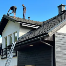 Dwóch dekarzy na dachu domu z ciemną dachówką, jeden wierci w dachu, drugi nadzoruje. Widoczna drabina oparta o ścianę budynku i błękitne niebo.