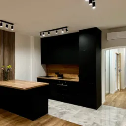 Nowoczesna kuchnia z czarnymi szafkami, drewnianym blatem i panelem dekoracyjnym na ścianie, oświetlona reflektorami sufitowymi, widok na przedpokój z drewnianą podłogą.