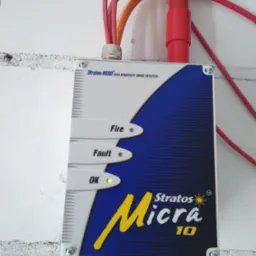 Zamontowany na ścianie detektor dymu Stratos-HSSD Micra 10 z sygnalizacją stanu: OK (zielona dioda), Fault i Fire, z widocznymi kablami i rurkami instalacyjnymi.