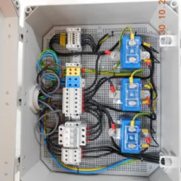 Otwarta szara skrzynka elektryczna z zamontowanymi bezpiecznikami, listwami zaciskowymi i przewodami o różnych kolorach, widoczne oznaczenia na przewodach i elementach, perforowane dno skrzynki.