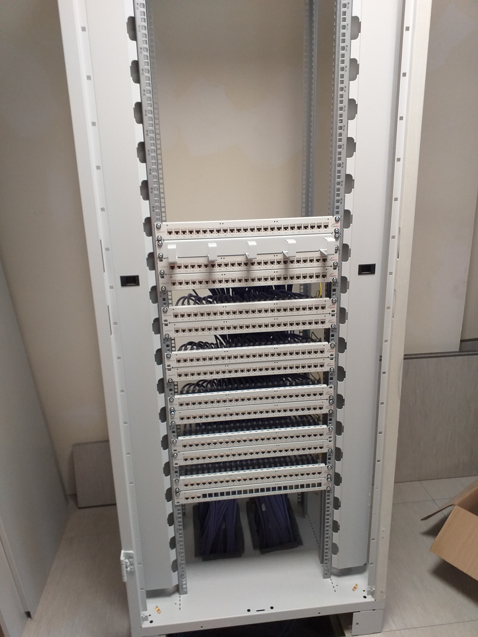 Wnętrze szafy rack z panelami krosowniczymi i uporządkowanymi kablami patchcord, widoczne mocowania i elementy konstrukcyjne szafy. Kable w dolnej części szafy spięte w wiązki.