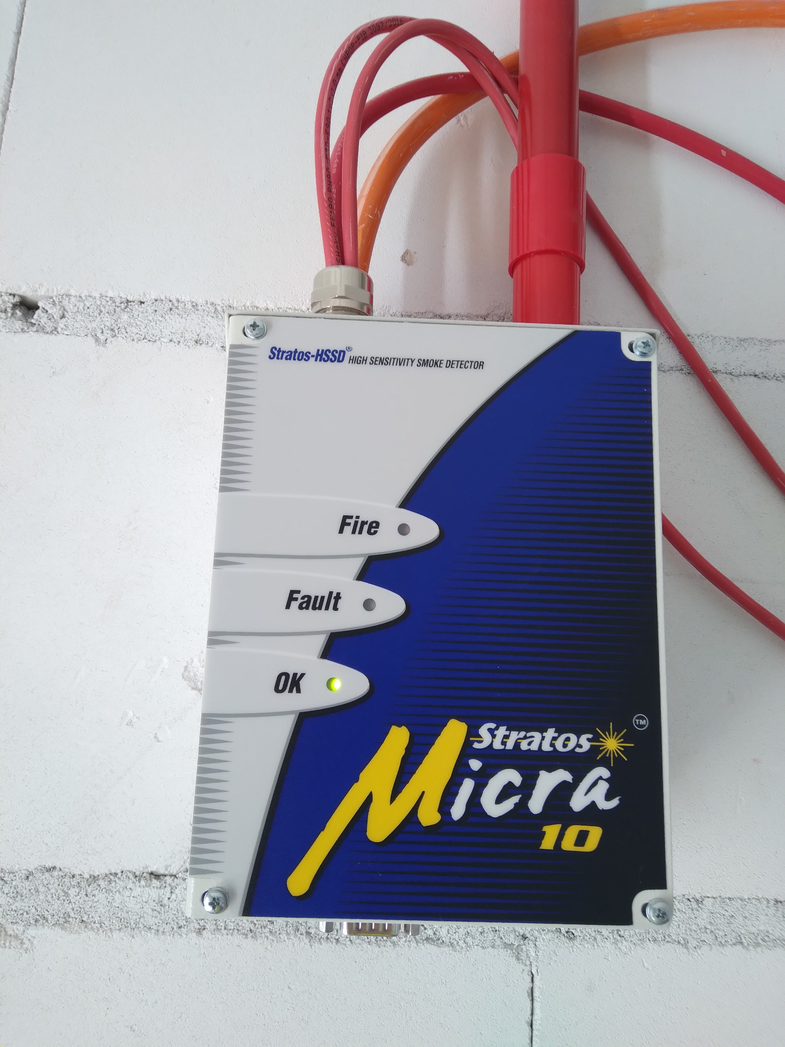 Zamontowany na ścianie detektor dymu Stratos-HSSD Micra 10 z sygnalizacją stanu: OK (zielona dioda), Fault i Fire, z widocznymi kablami i rurkami instalacyjnymi.