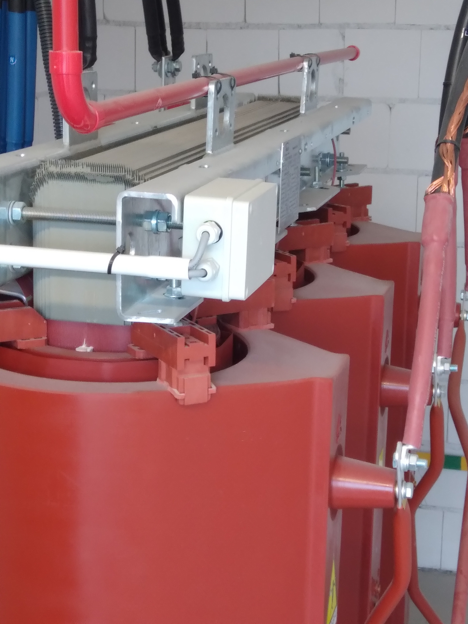 Widok z bliska na transformator energetyczny z czerwonymi obudowami, białymi elementami montażowymi i grubymi kablami, umieszczony w pomieszczeniu z białymi ścianami z bloków.