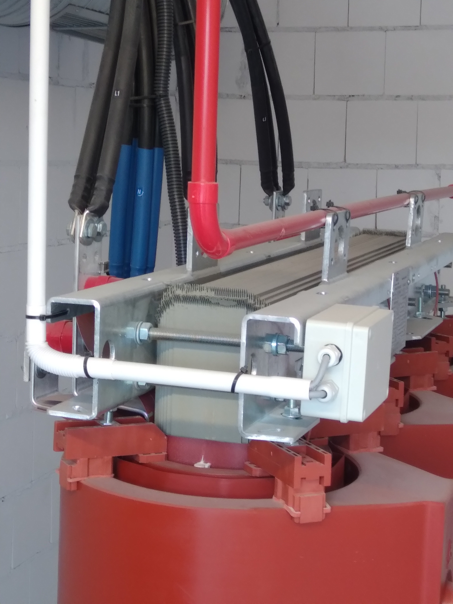 Fragment transformatora z widocznymi przewodami elektrycznymi w kolorach czarnym, czerwonym i niebieskim, zamocowanymi do metalowej konstrukcji na tle ściany z białych bloczków.