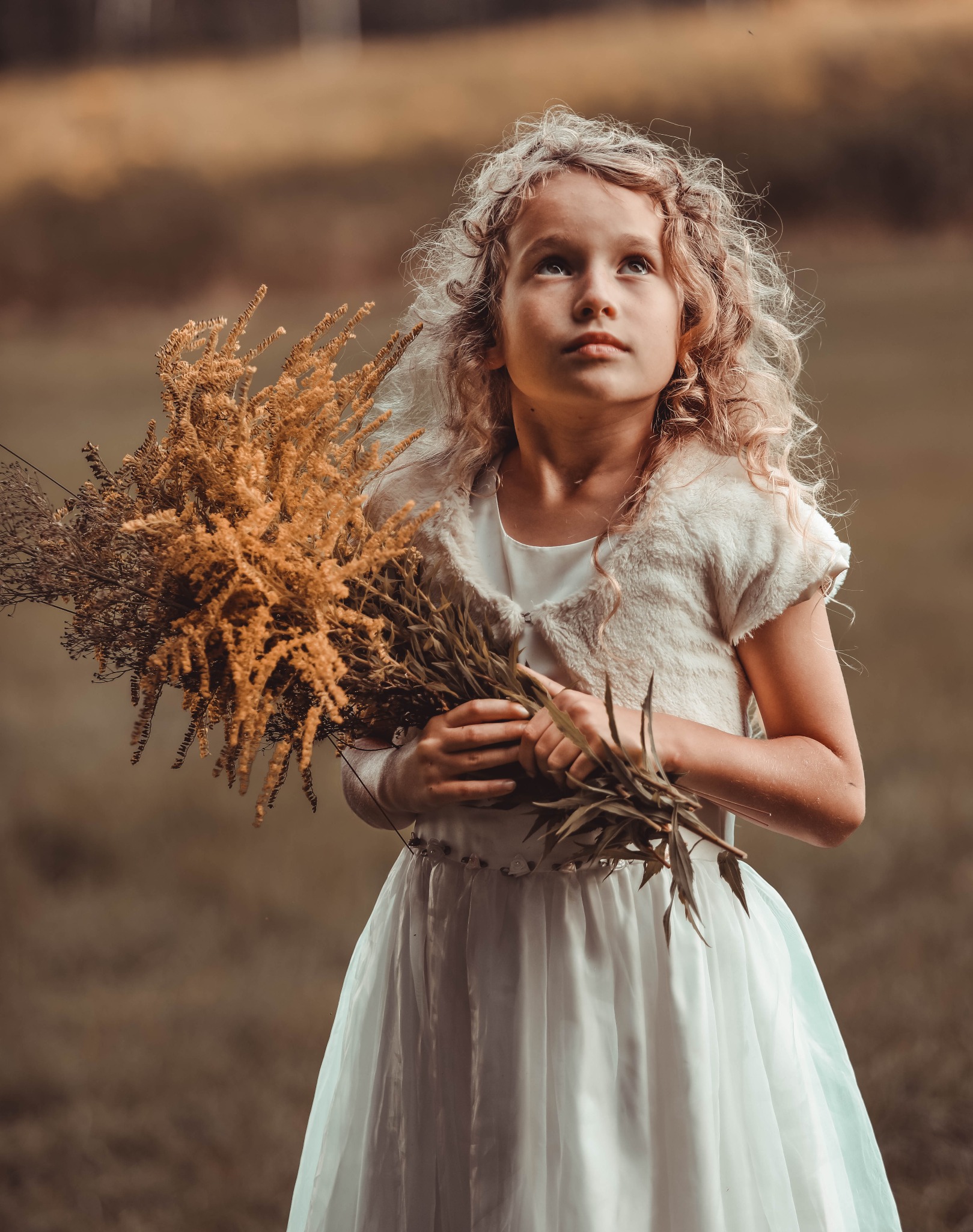 Dziewczynka z blond lokami w białej sukience i futrzanym bolerku, trzymająca bukiet suszonych traw, spogląda w górę na tle rozmytej zieleni.