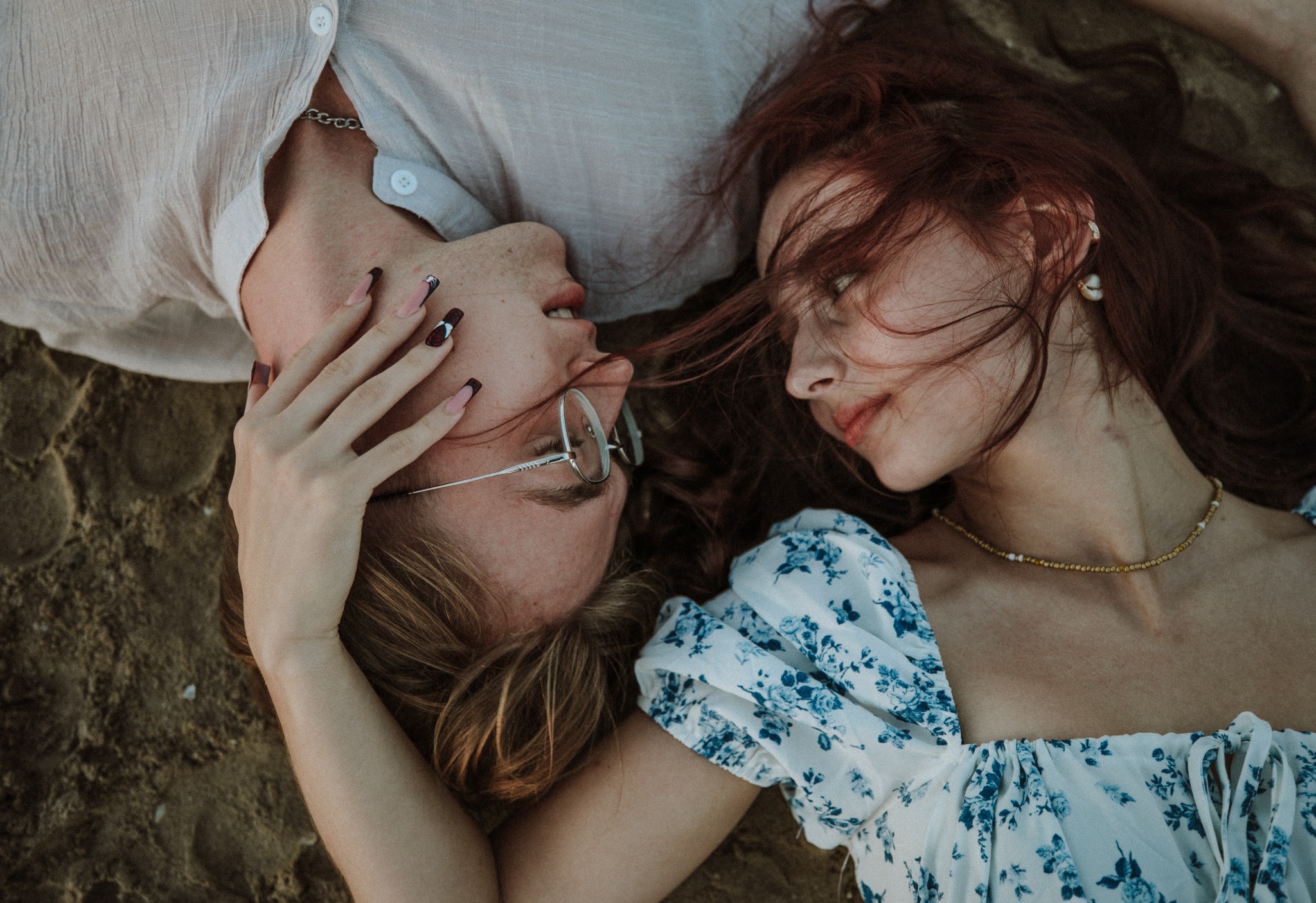 Para leżąca na piasku, wzajemnie wpatrzona w siebie; ona w kwiecistej sukience, on w okularach, z dłonią na jego twarzy, podkreślona artystycznym manicure.