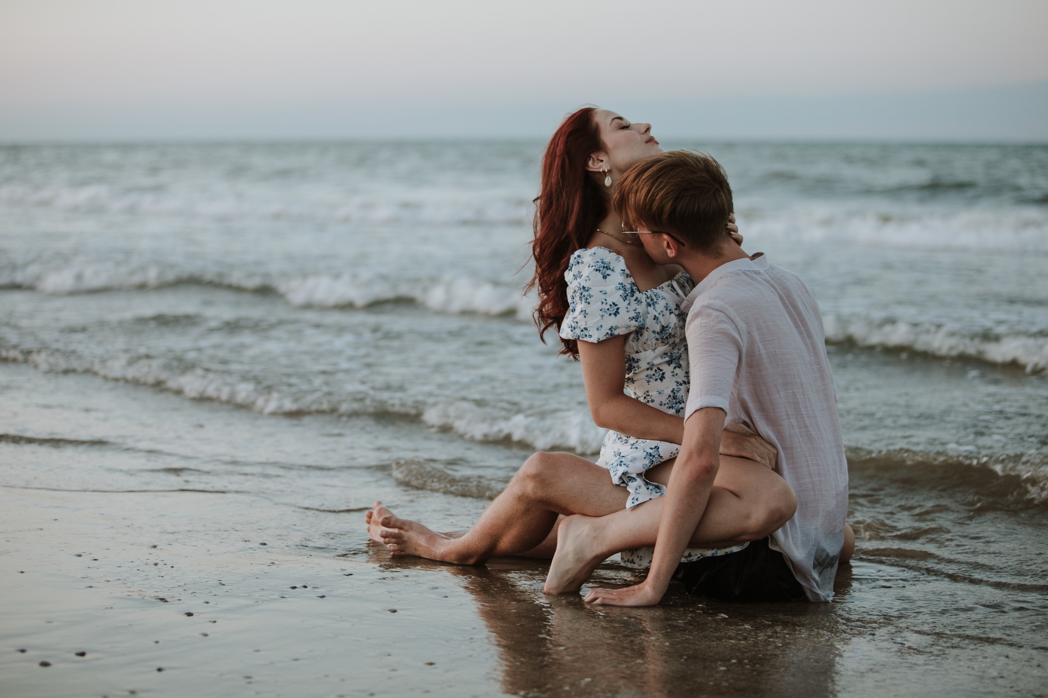 Para zakochanych siedzi na mokrym piasku na plaży, kobieta z rudymi włosami w kwiecistej sukience odchyla głowę do tyłu, mężczyzna w białej koszuli obejmuje ją.