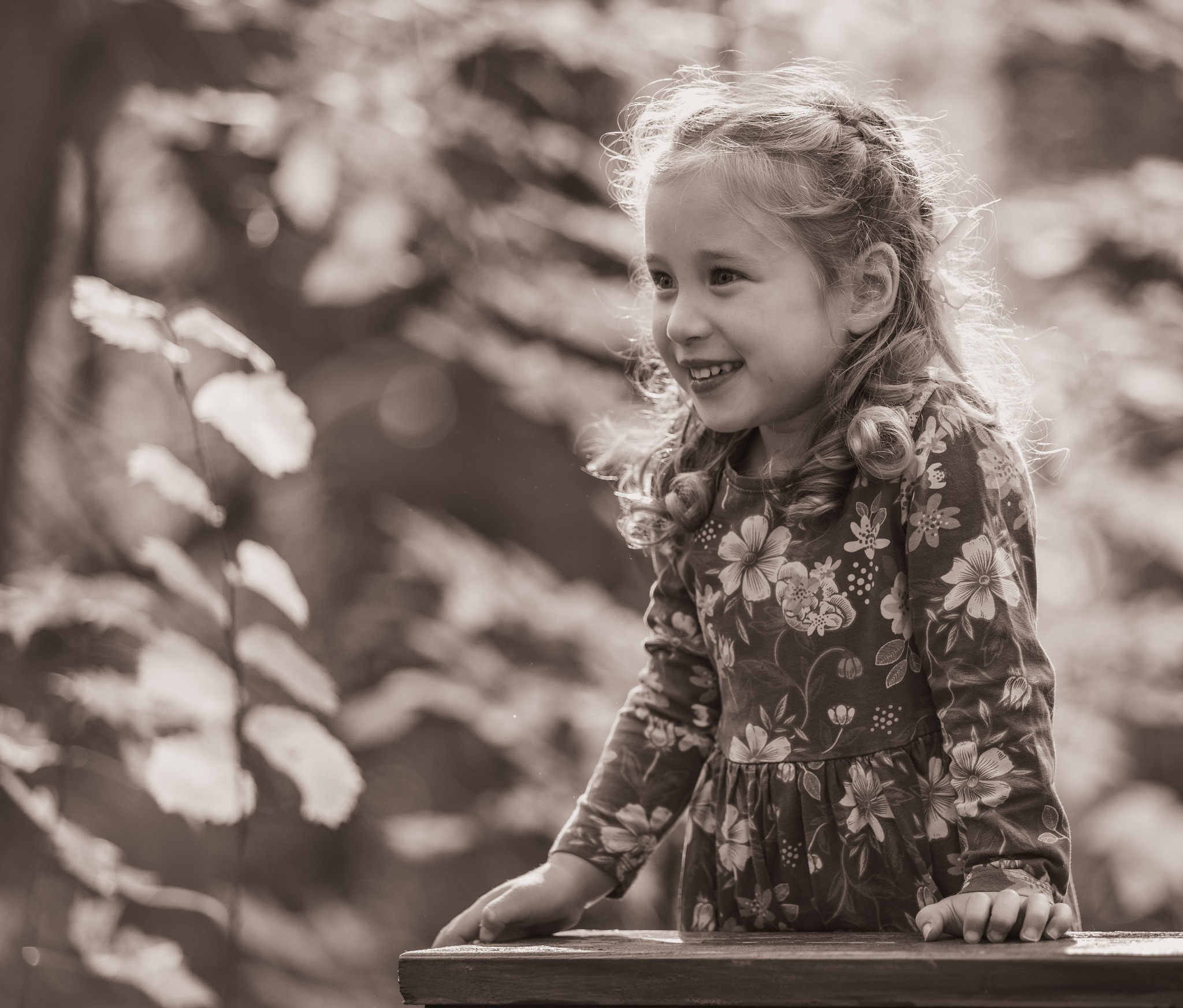 Portret uśmiechniętej dziewczynki w sukience w kwiaty, z warkoczem i podświetlonymi włosami, na tle rozmytej zieleni.