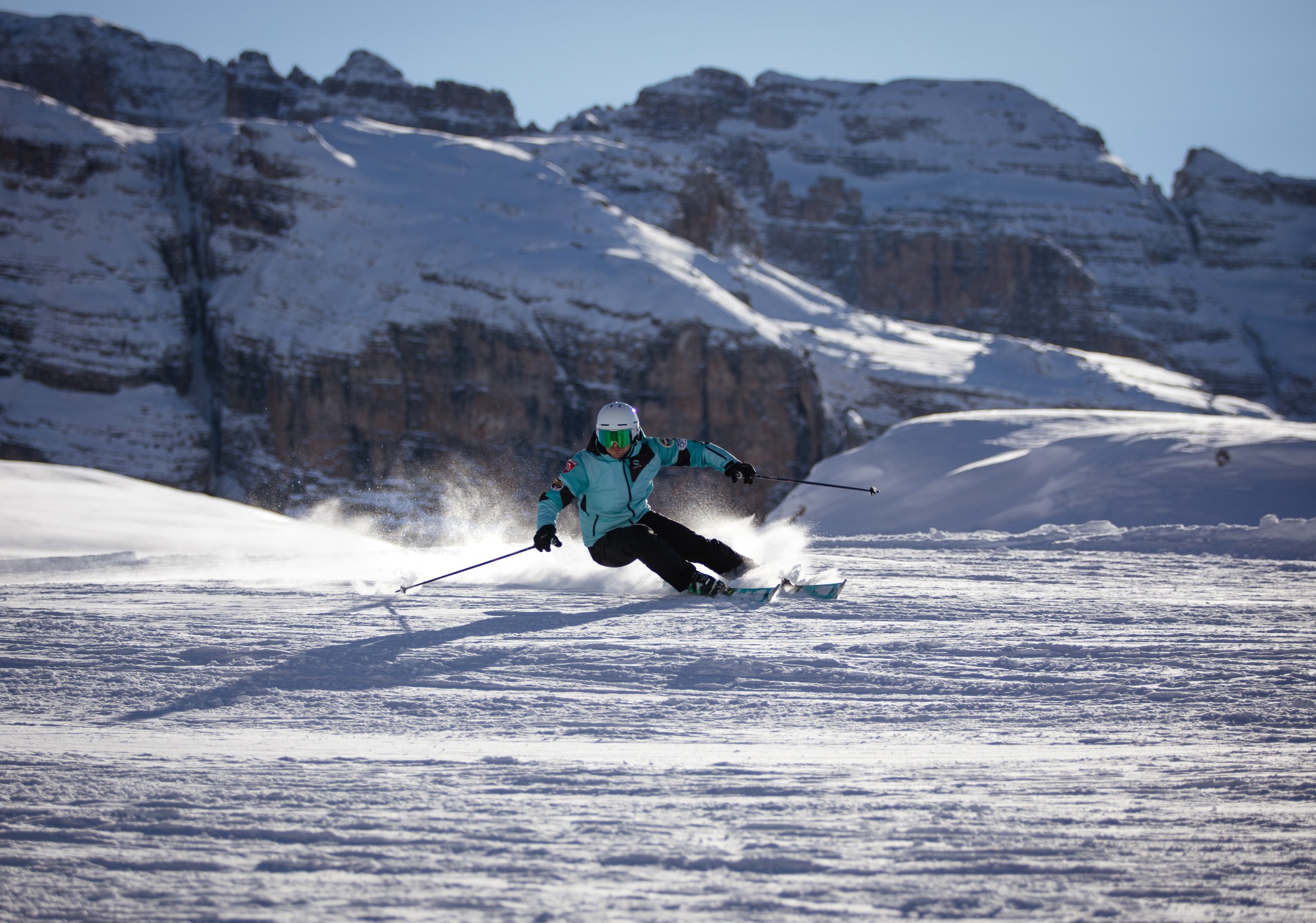 Narciarz w turkusowej kurtce i białym kasku dynamicznie zjeżdża po ośnieżonym stoku, w tle majestatyczne, ośnieżone góry i błękitne niebo.