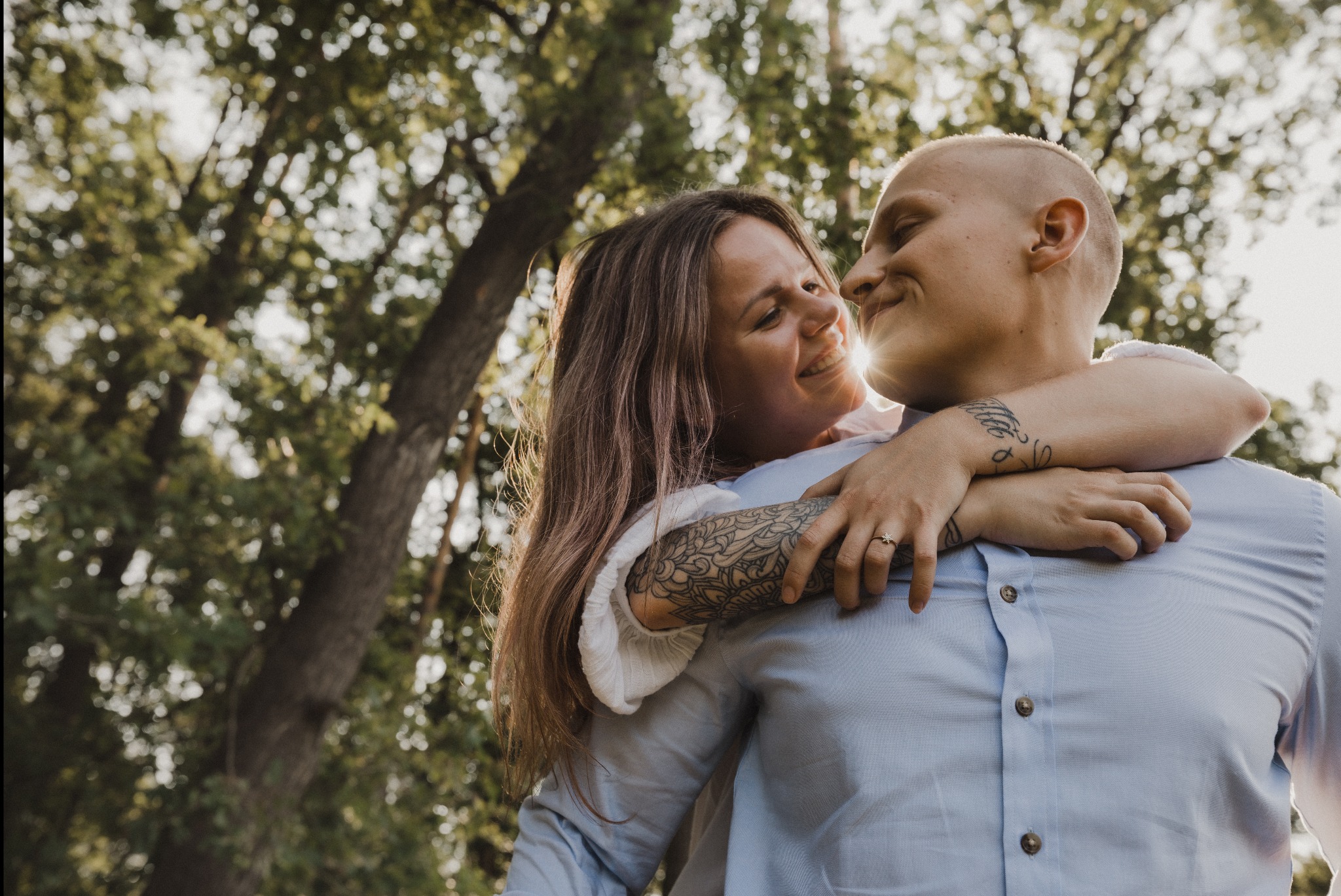 Uśmiechnięta para narzeczonych w objęciach na tle drzew, widoczne tatuaże na ramionach, delikatne światło słoneczne.