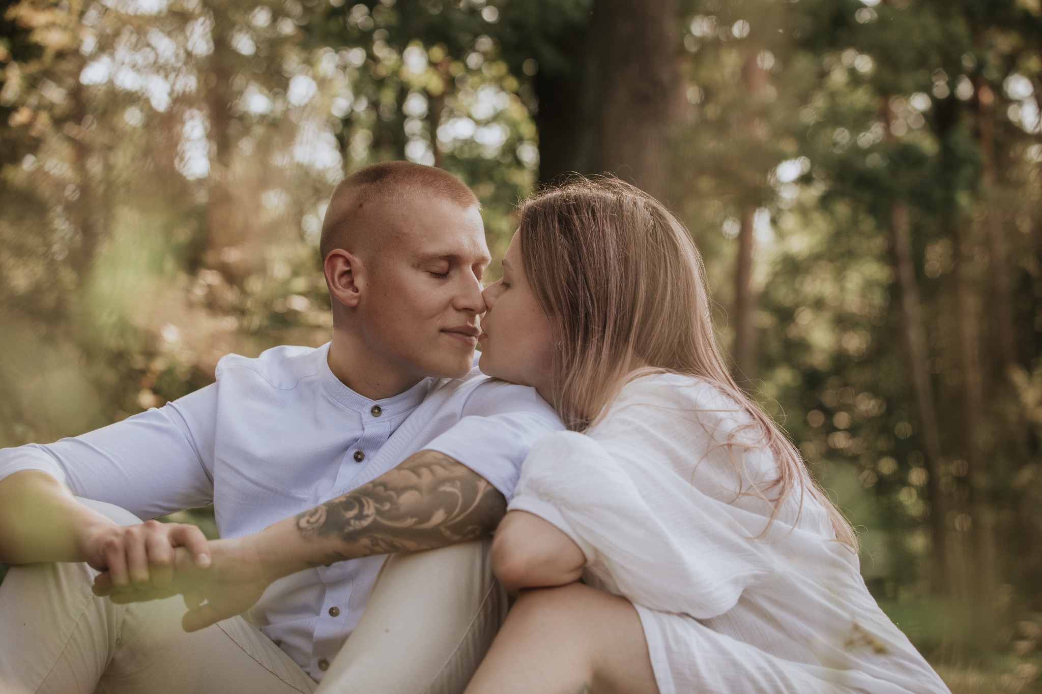 Para zakochanych siedzi w lesie, dotykając się nosami, mężczyzna ma tatuaż na ramieniu.