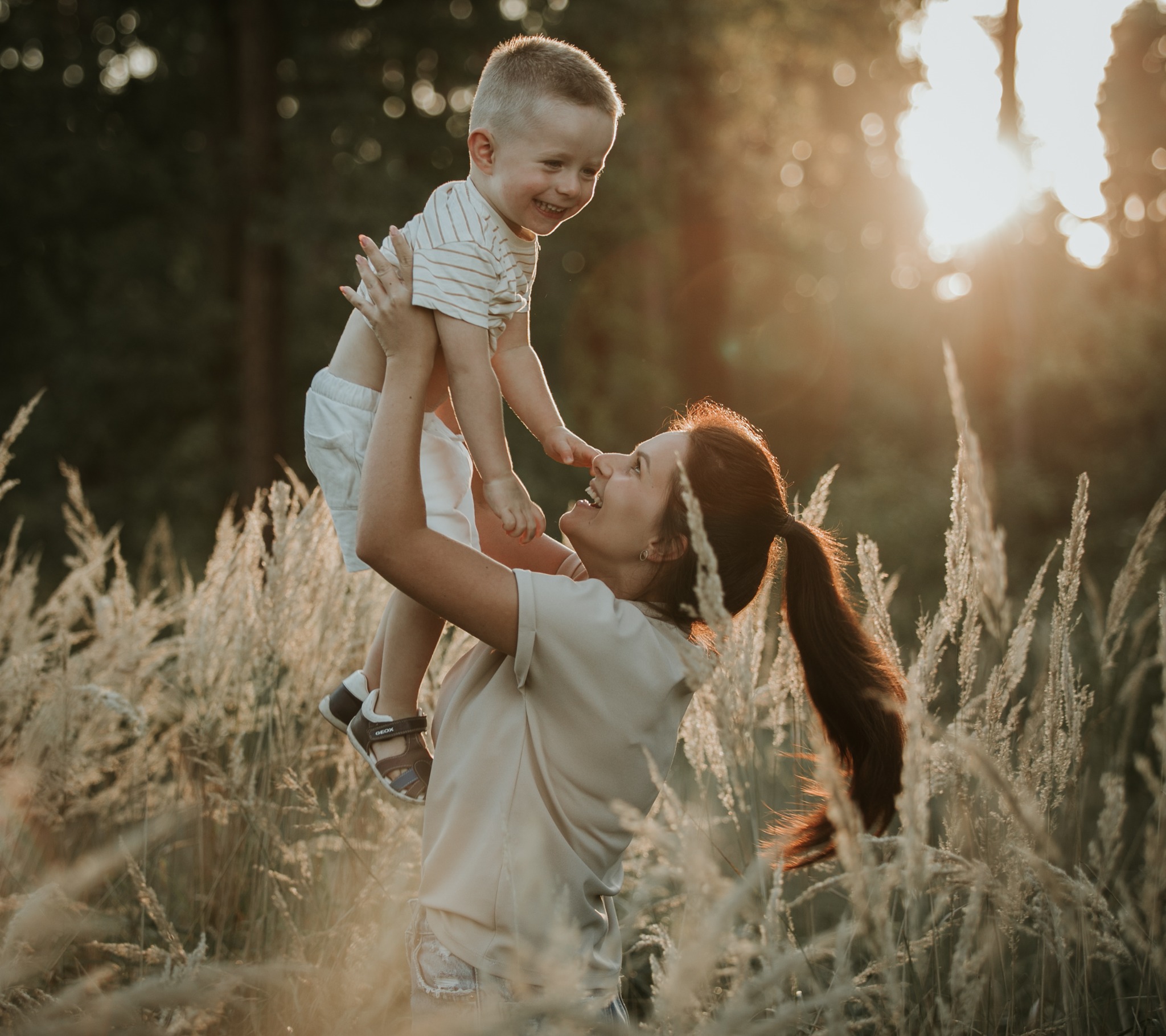 Uśmiechnięta matka podnosi w górę roześmianego synka na tle łąki w promieniach zachodzącego słońca.