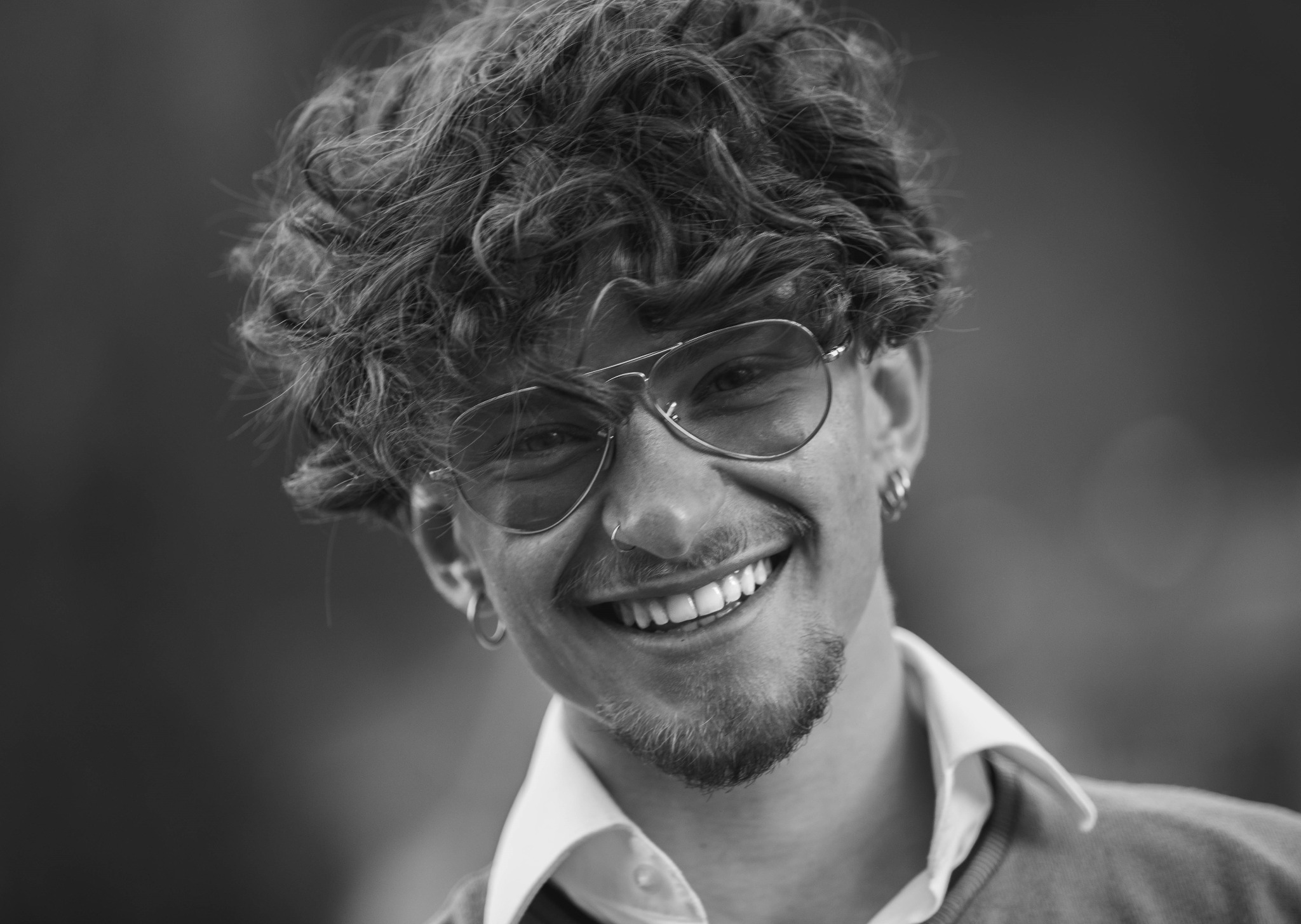 Czarno-biały portret uśmiechniętego mężczyzny z kręconymi włosami, noszącego okulary typu aviator, kolczyki i pierścień w nosie.