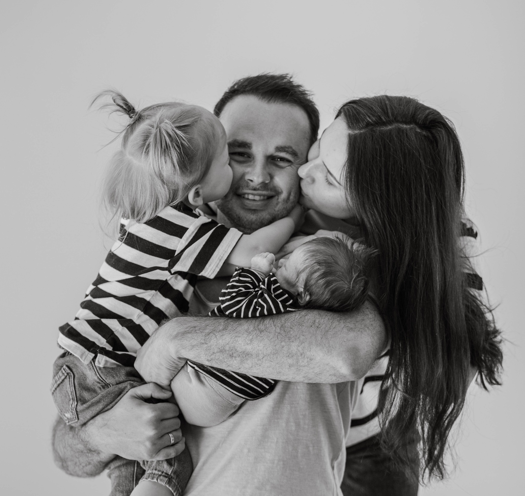 Czarno-białe zdjęcie uśmiechniętego ojca, którego z jednej strony całuje mała dziewczynka w bluzce w paski, a z drugiej strony mama z długimi włosami; na rękach ojca śpi niemowlę również w bluzce...