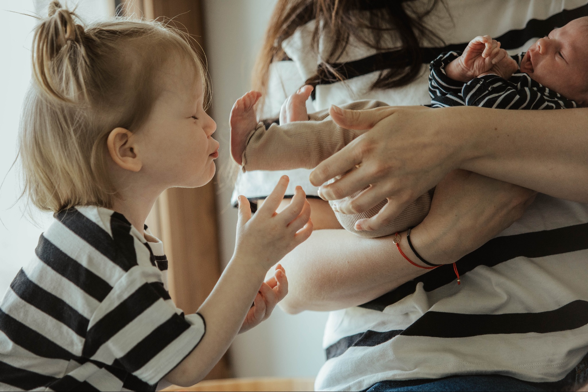 Ujęcie z Gliwic przedstawia starszą siostrę w czarno-białej koszulce w paski, która z zaciekawieniem przygląda się śpiącemu noworodkowi trzymanemu przez matkę.