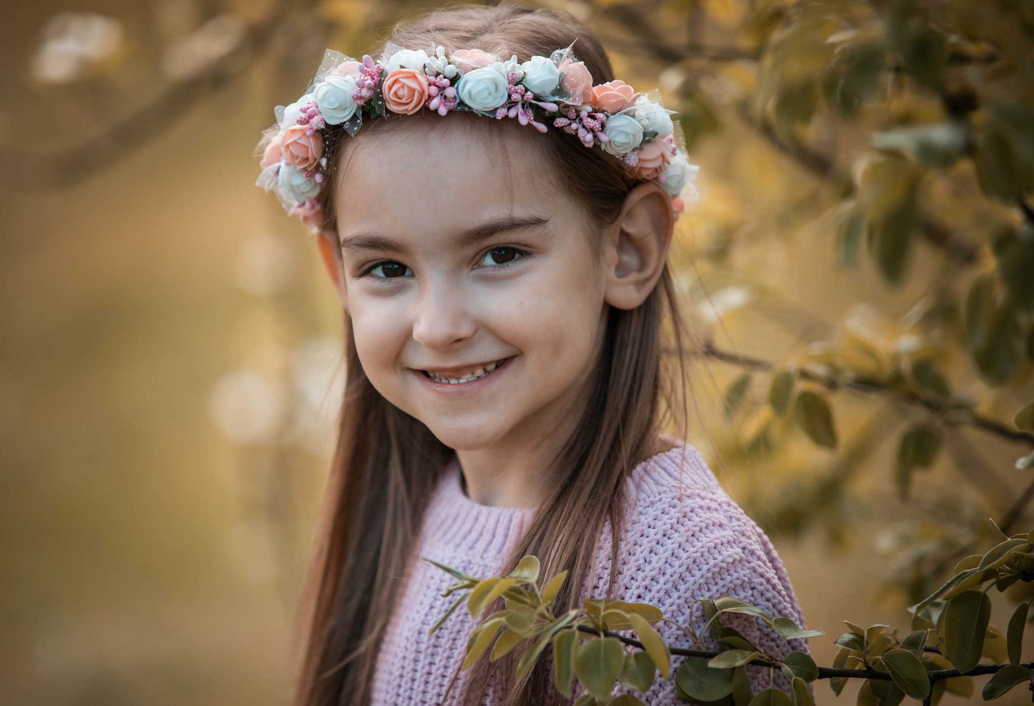 Portret uśmiechniętej dziewczynki w wianku z różowych i białych kwiatów, ubrana w liliowy sweter, na tle jesiennych liści.