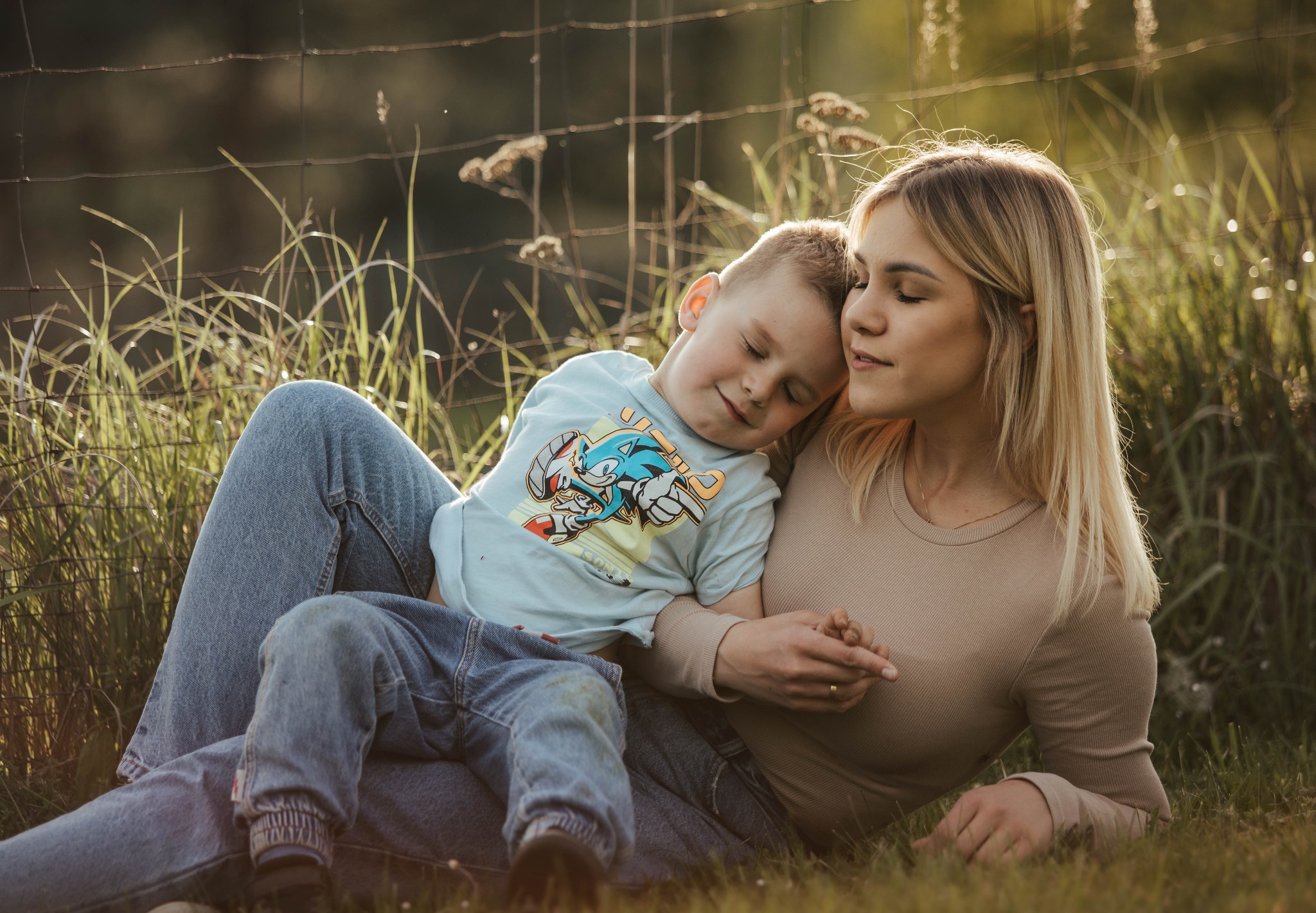 Matka z synem leżą na trawie, chłopiec śpi oparty o nią, oboje ubrani w dżinsy, w tle siatka ogrodzeniowa.