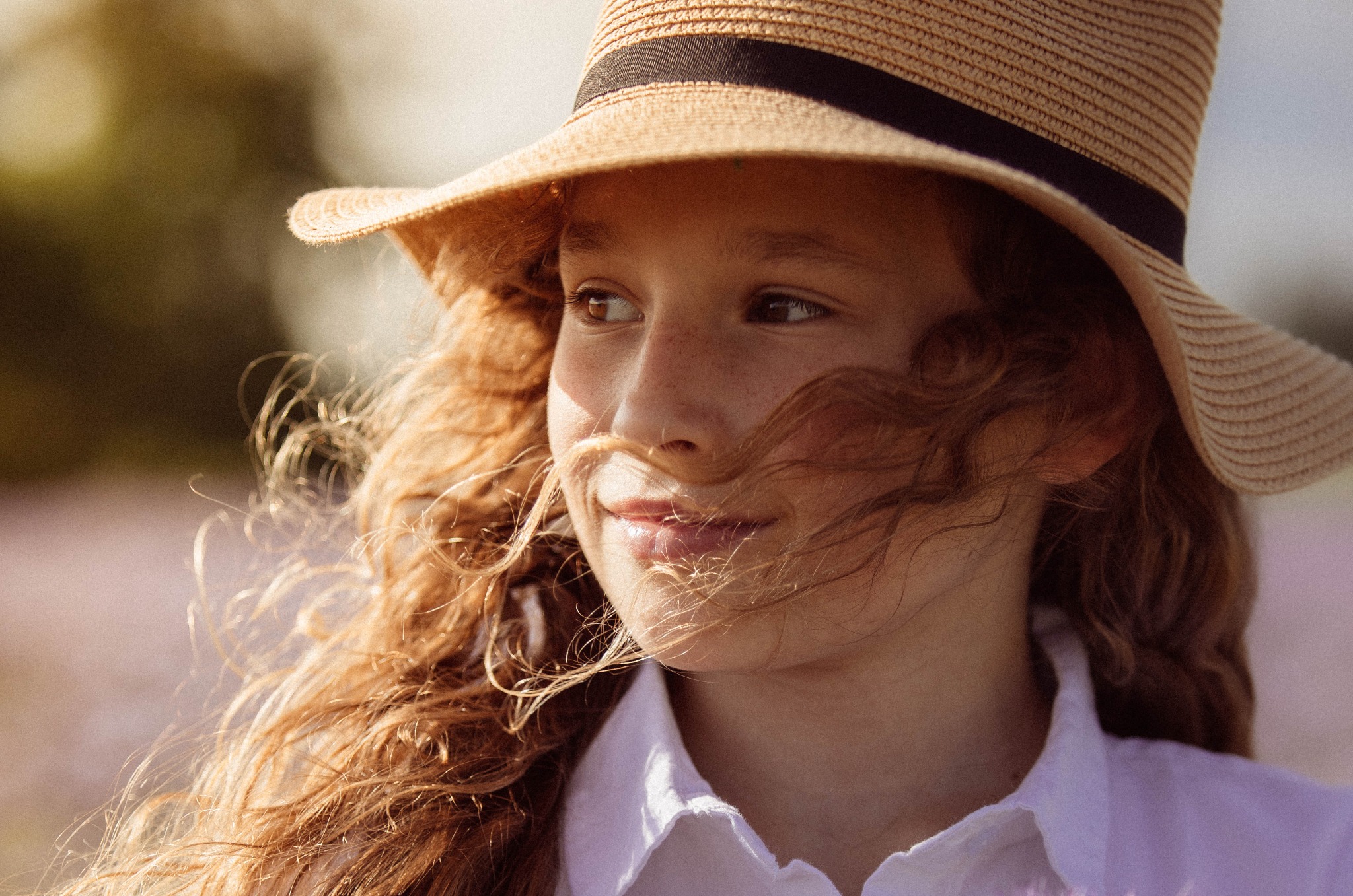 Portret młodej dziewczyny z rudymi kręconymi włosami i piegami, w słomkowym kapeluszu, spoglądającej w bok na tle rozmytego pola lawendy.